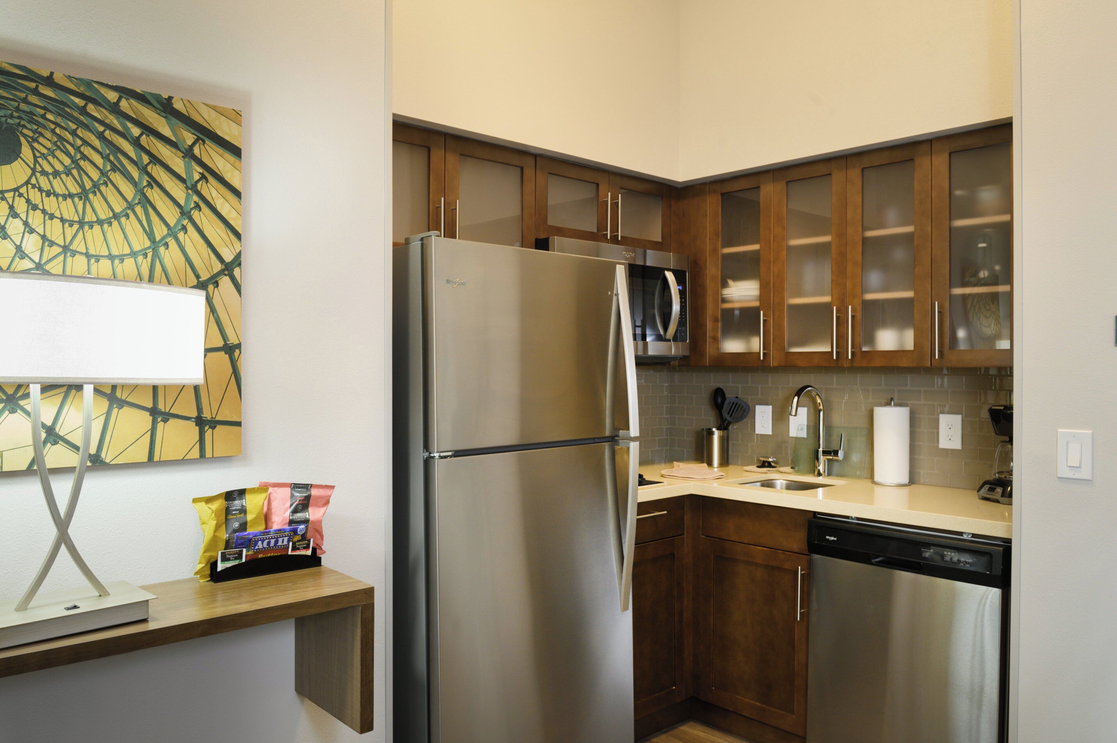 A compact kitchen with a refrigerator, microwave, and dishwasher. There is a wooden cabinet with a glass door, a countertop with a sink, and a wall-mounted light fixture.