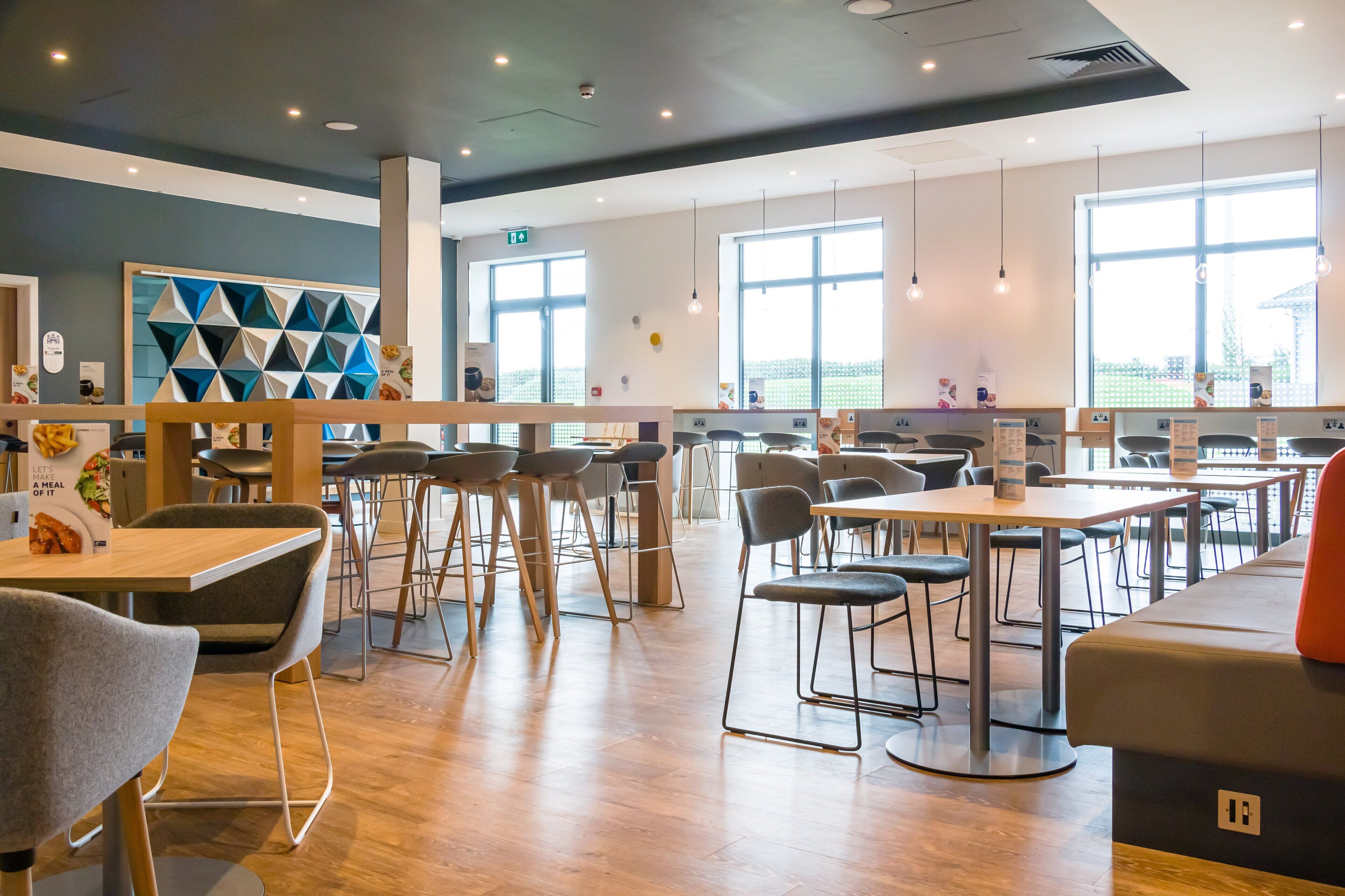A modern dining area with wooden tables and chairs, large windows, and a geometric wall decoration.