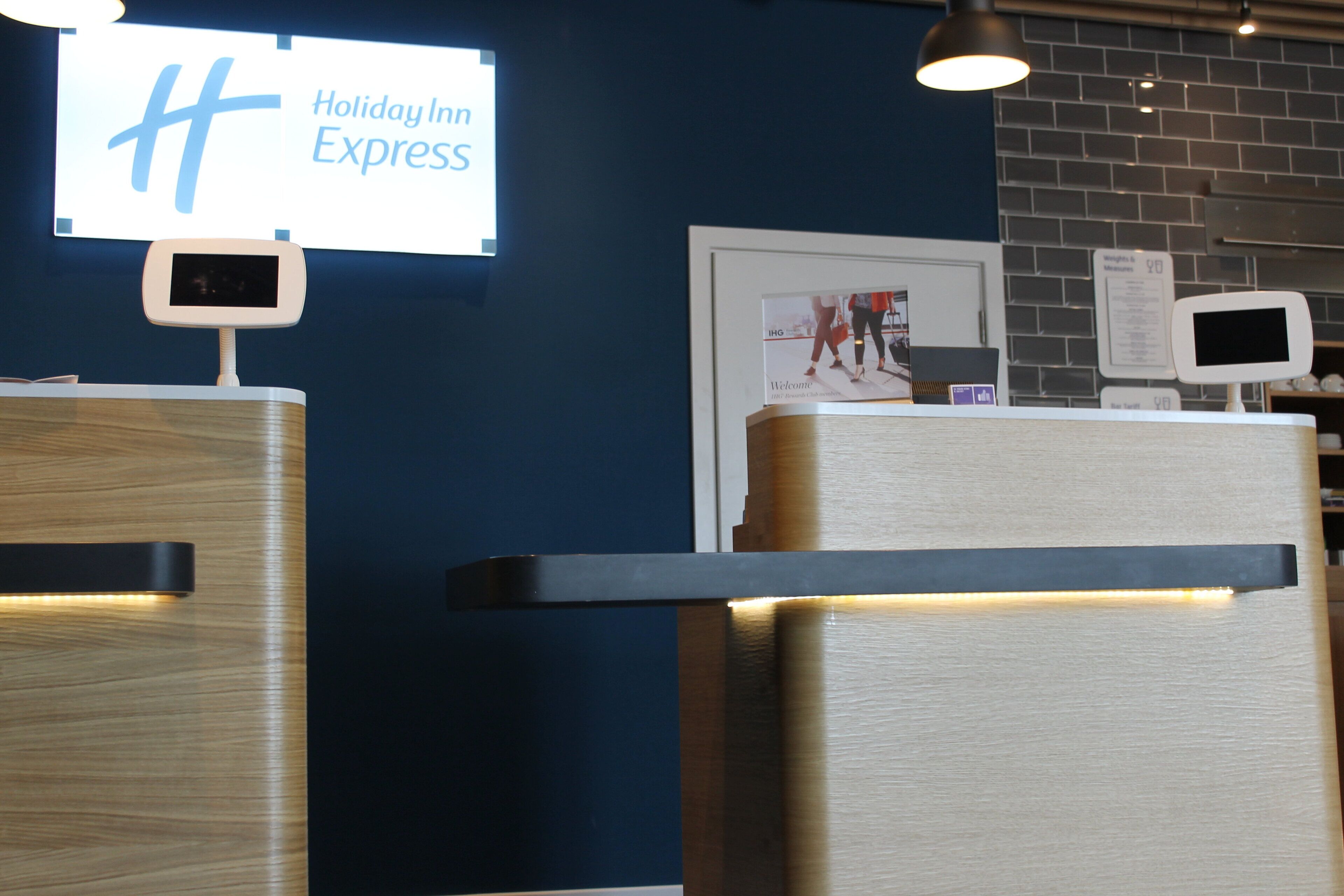 A Holiday Inn Express reception desk with a digital display, framed welcome poster, and a brick wall background.
