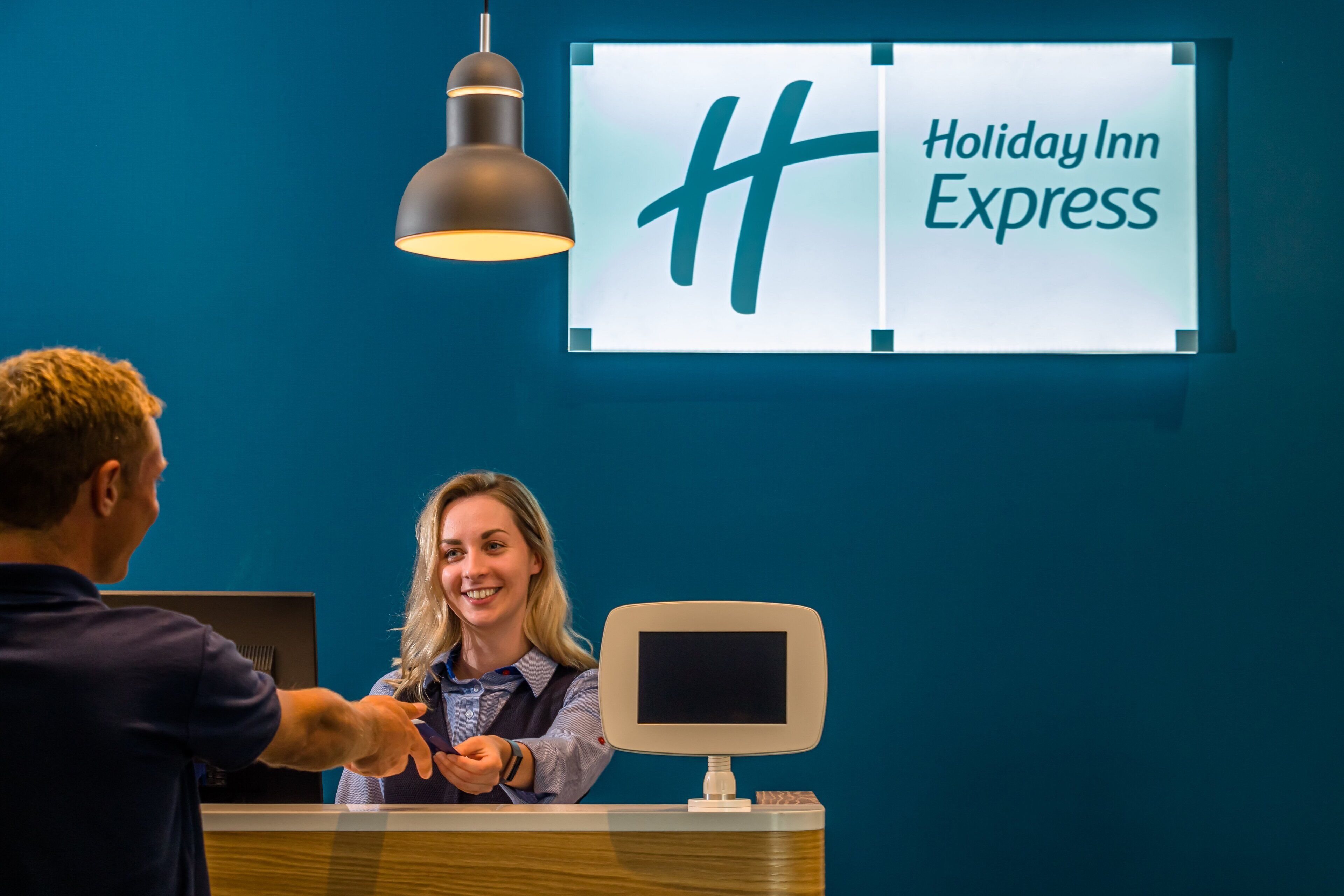 A Holiday Inn Express reception desk with a large sign above it, a desk lamp, and a computer monitor.