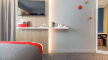 Une chambre d’hôtel dotée d’un téléviseur à écran plat fixé au mur, d’une petite étagère avec une bouilloire et des tasses, et d’une touche de couleur rouge sur le lit.