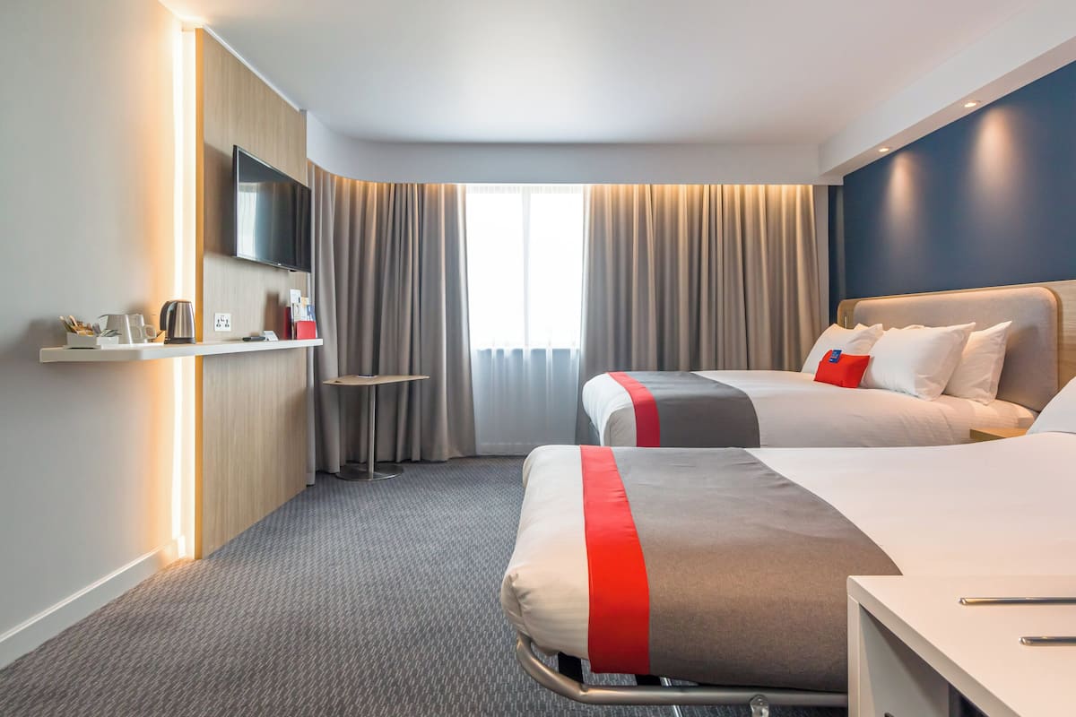 Habitación de hotel con dos camas, televisor, escritorio con lámpara y ventana con cortinas.