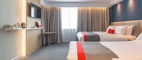 A hotel room with two beds, a TV, a desk with a lamp, and a window with curtains.