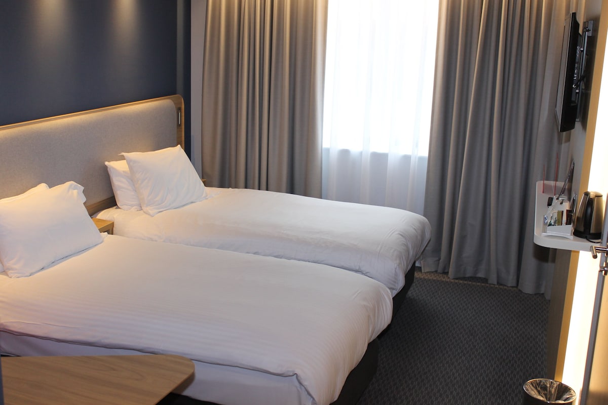 Een hotelkamer met twee bedden, een nachtkastje, een aan de muur bevestigde televisie en een raam met gordijnen.