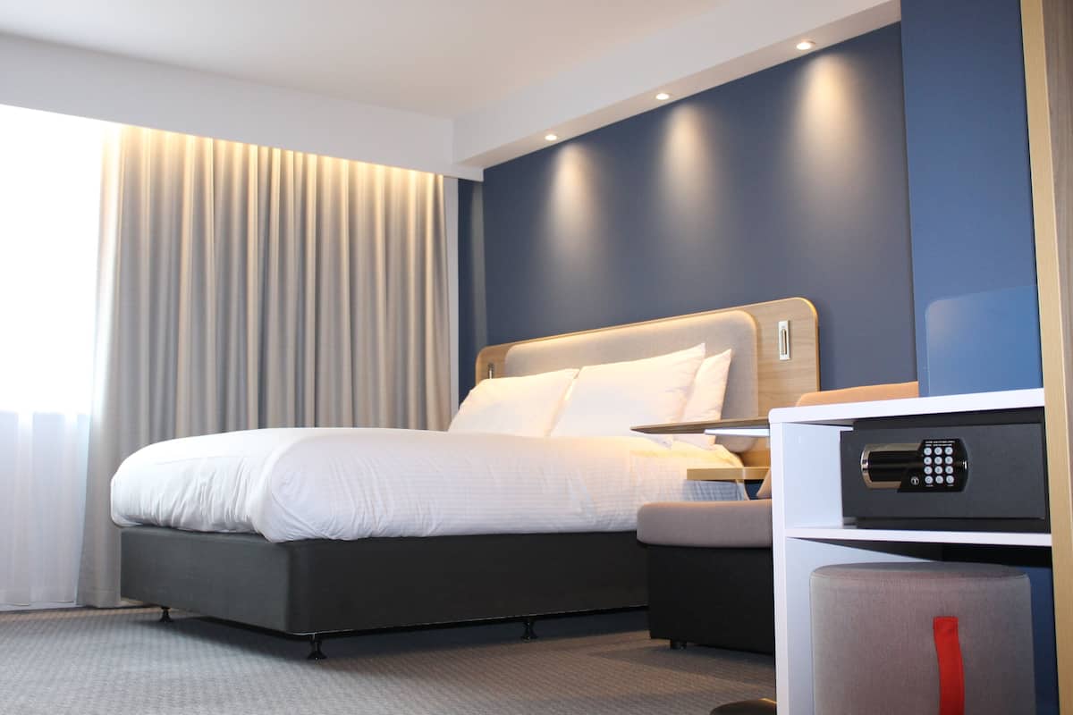 Habitación de hotel moderna con una cama grande, un escritorio con panel de control y vistas a la ciudad por la ventana.
