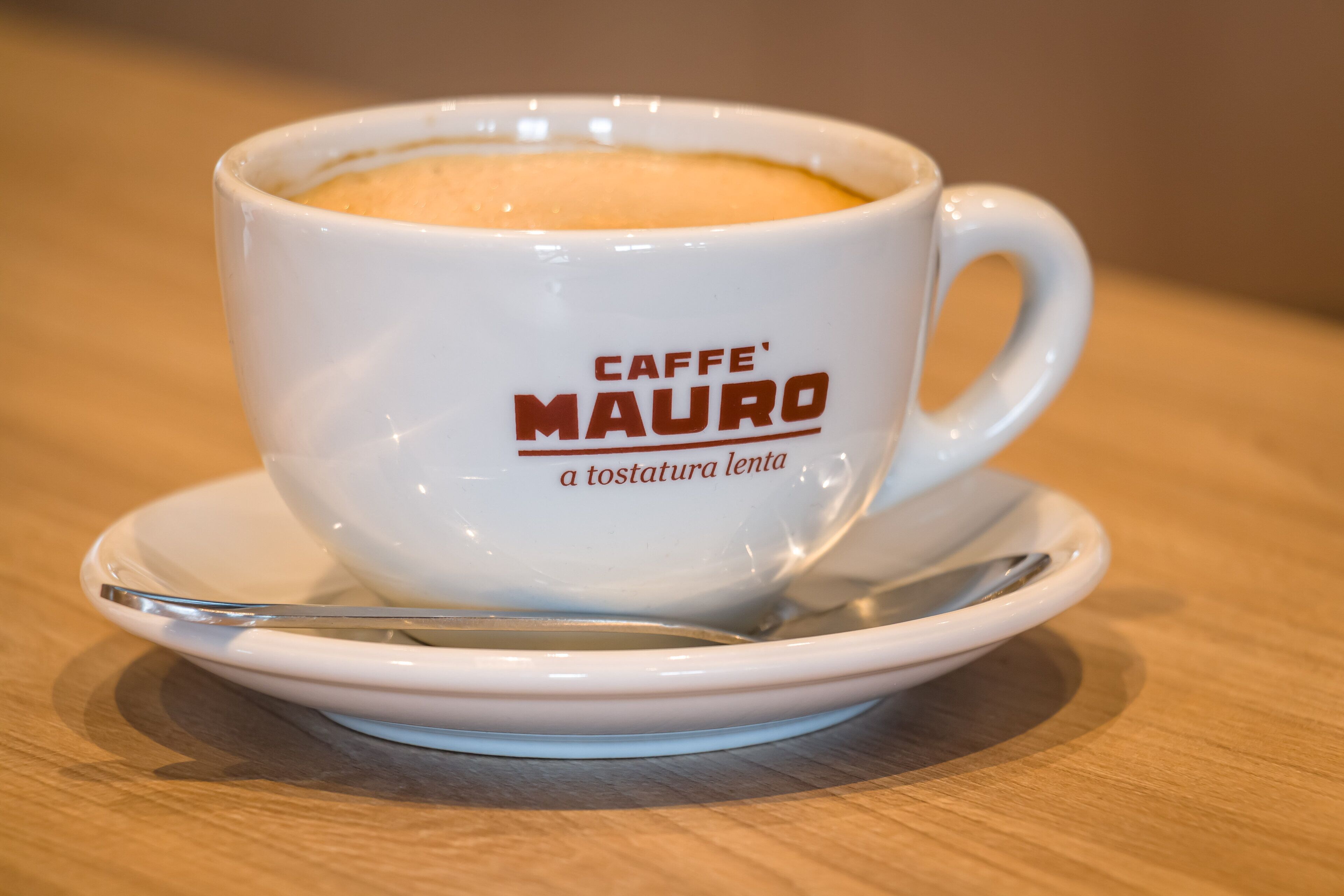 Une tasse blanche portant les inscriptions « CAFFE' MAURO » et « a tostatura lenta », posée sur une soucoupe avec une cuillère.