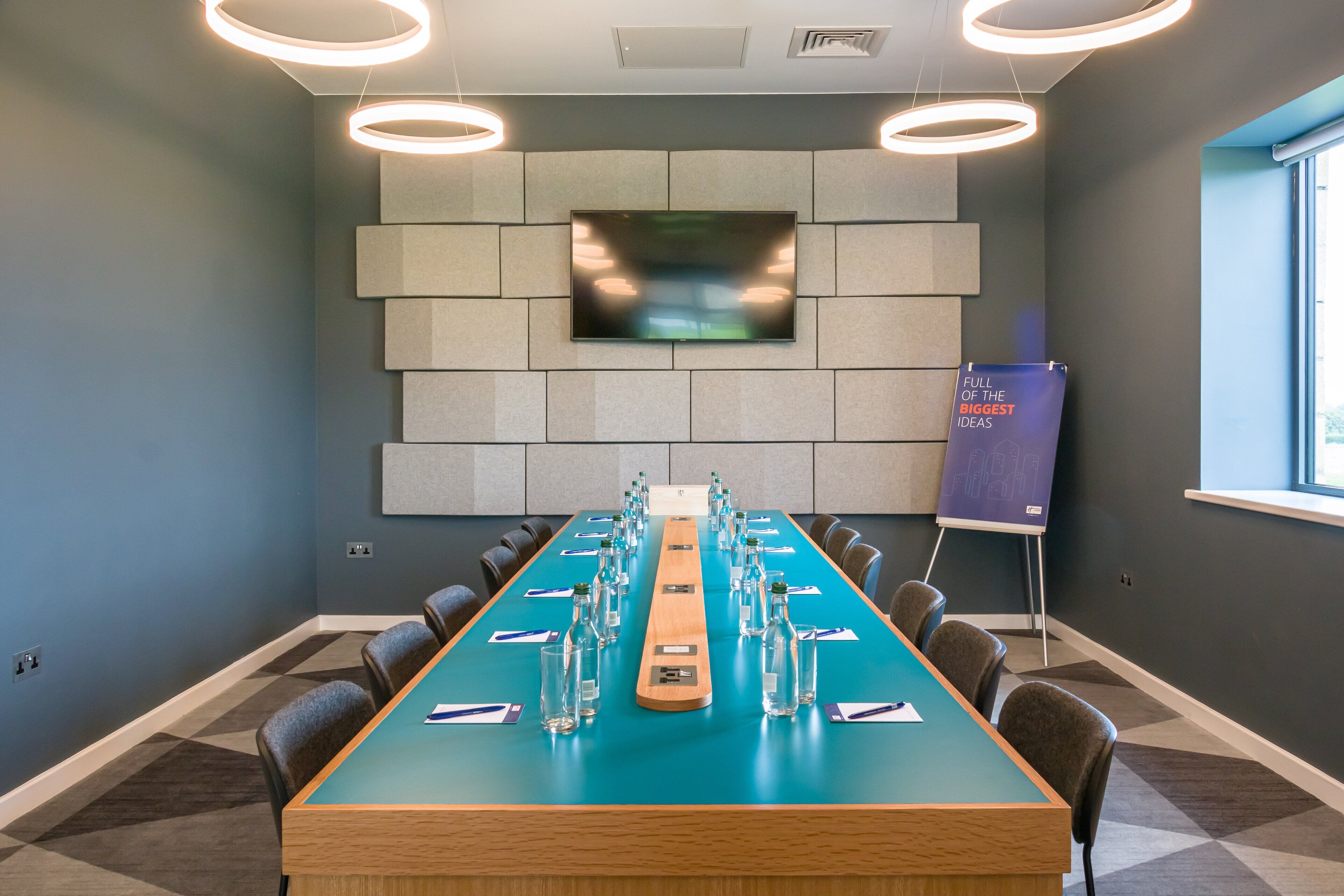 Un salón de conferencias con una larga mesa, sillas, un televisor y un atril con un letrero.
