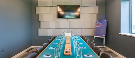 Un salón de conferencias con una larga mesa, sillas, un televisor y un atril con un letrero.