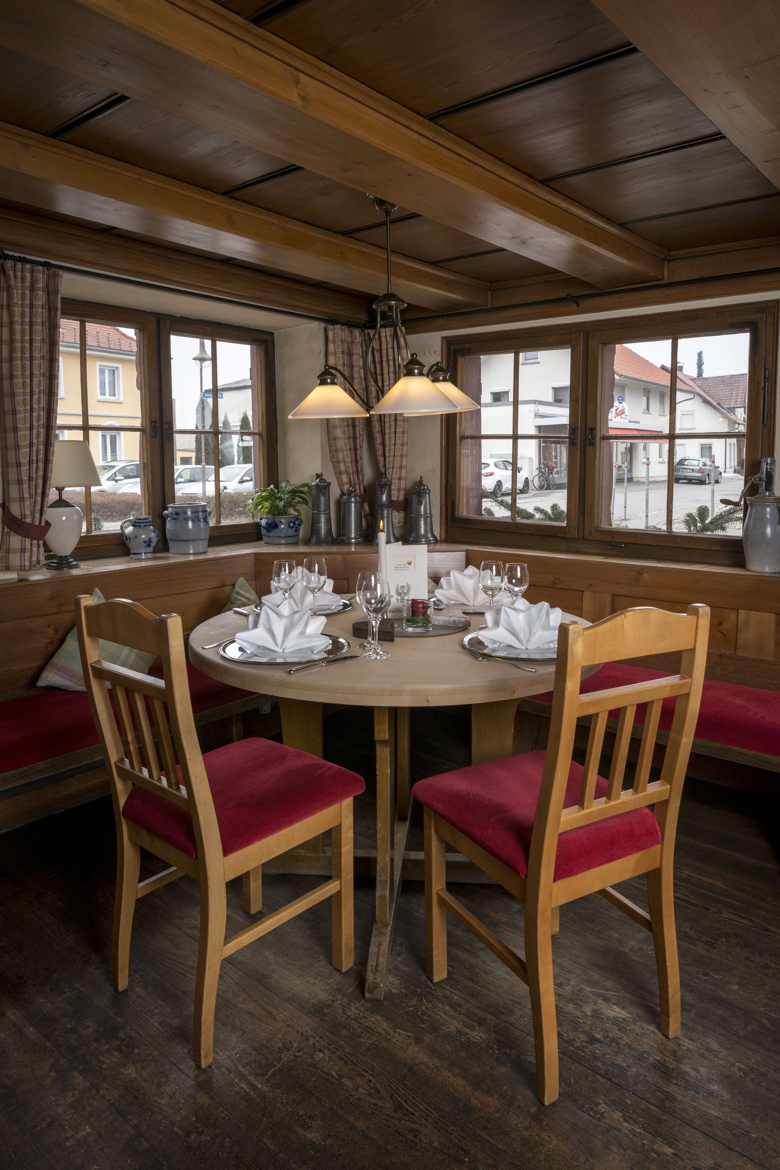 A wooden dining table set for four with red cushioned chairs, a chandelier, and a view of the street through large windows.