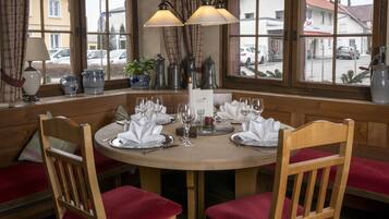 A wooden dining table set for four with red cushioned chairs, a chandelier, and a view of the street through large windows.