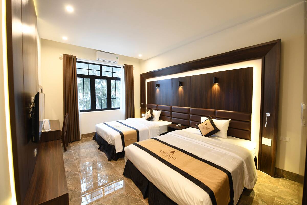 A hotel room with two beds, a wooden headboard, a desk, and a window with curtains.