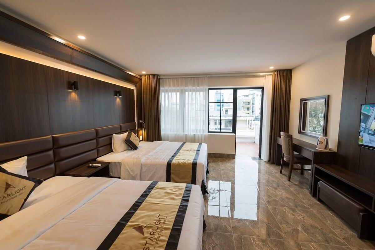 A hotel room with two beds, a desk, a TV, and a balcony with a view of buildings.