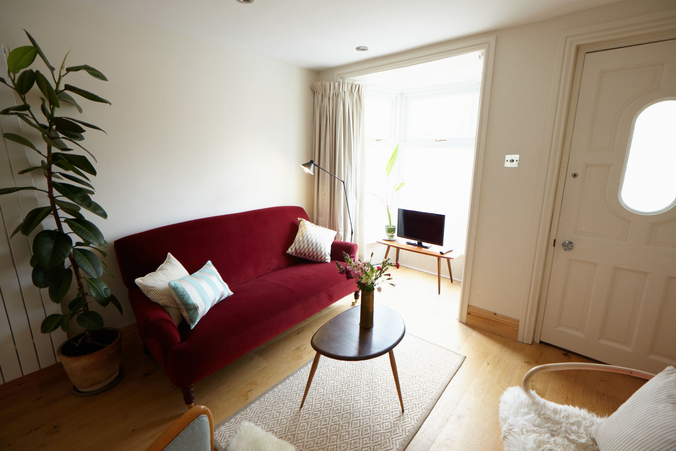 A living room with a red sofa, a small round table with a vase, a white furry chair, and a large potted plant.