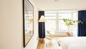 Een hotelkamer met een bed, een stoel, een vloerlamp, een raam met gordijnen en een lijst met een foto aan de muur.