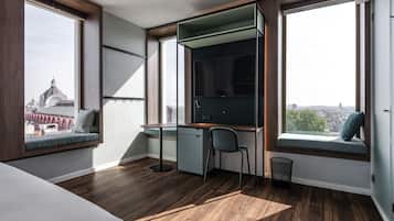 A modern bedroom with a desk, chair, and a view of a cityscape through large windows.