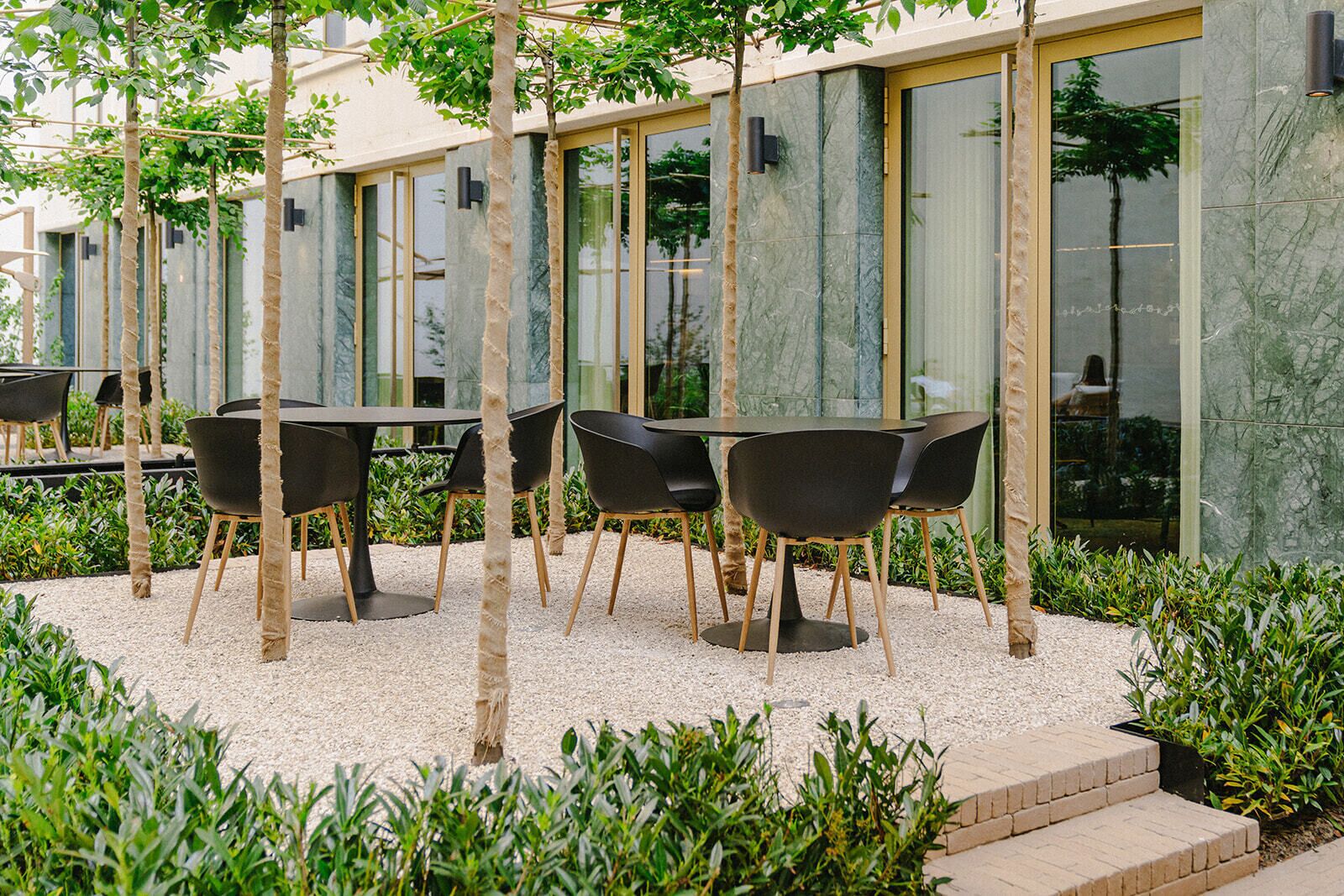 Un espacio exterior moderno con mesas y sillas, rodeado por un camino de grava y vegetación.
