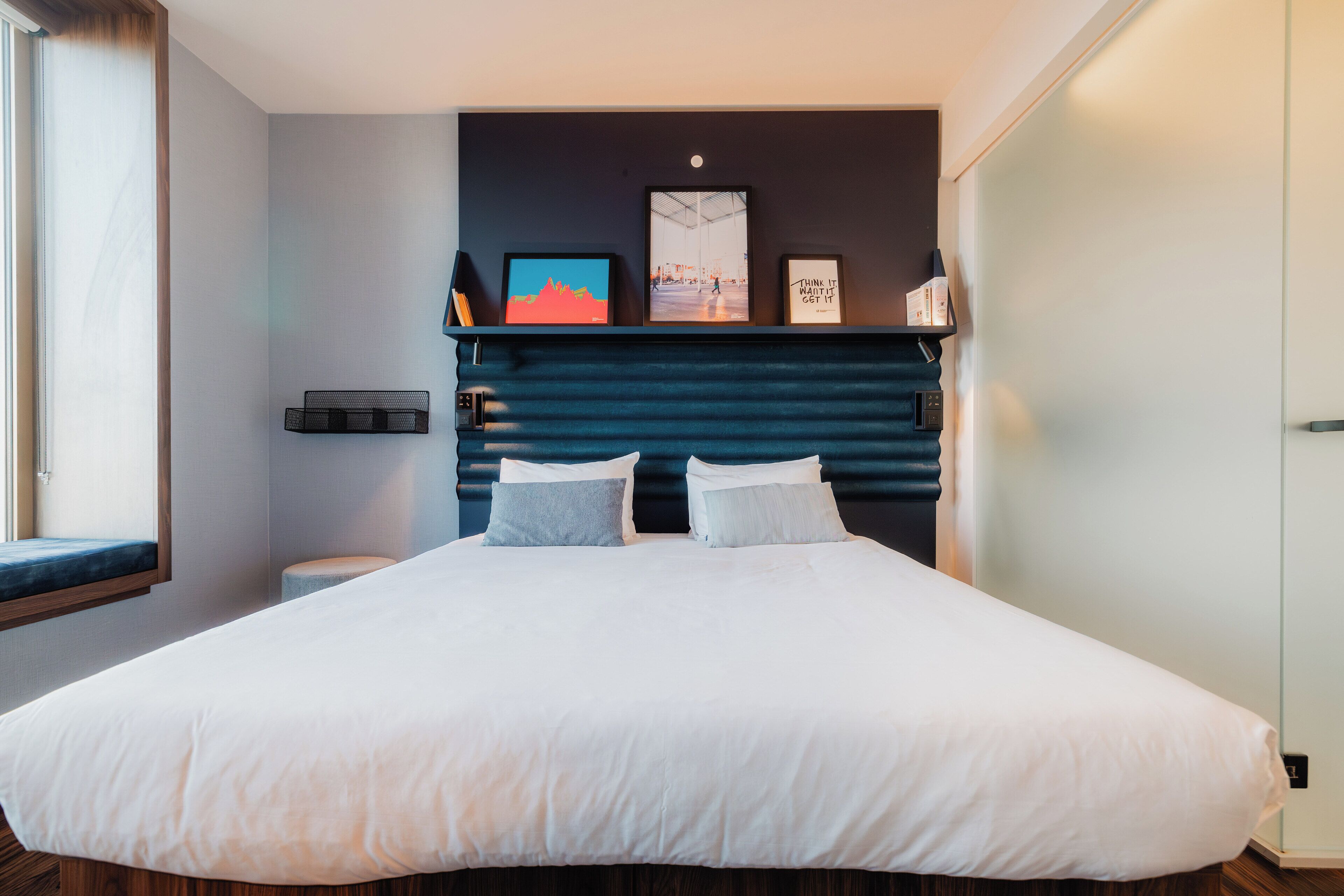 A modern hotel room with a large bed, a dark headboard, and a window with a view.