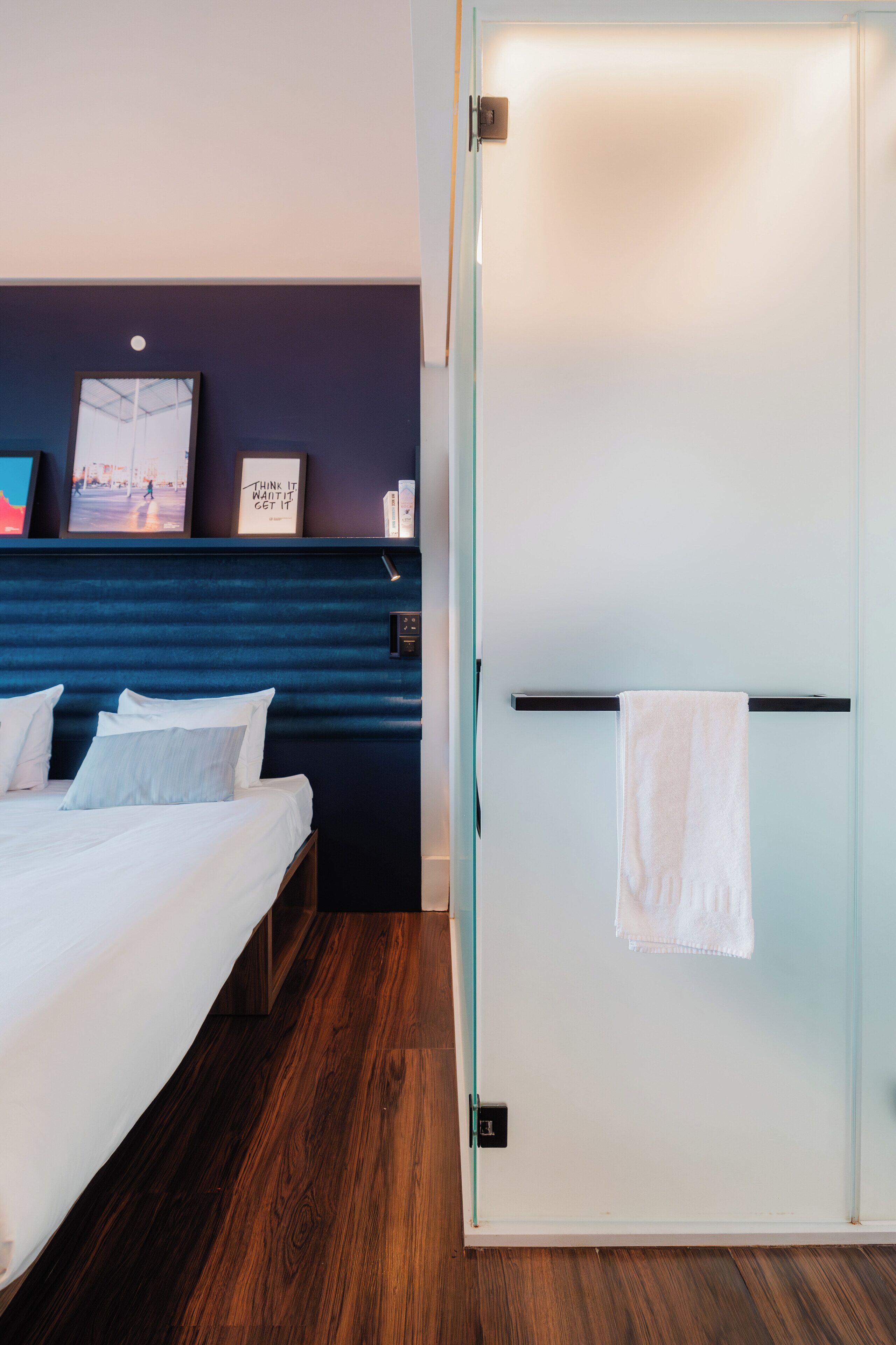 Una habitación de hotel con una cama, una mesita de noche, un estante con marcos y una puerta corredera con una toalla colgada.