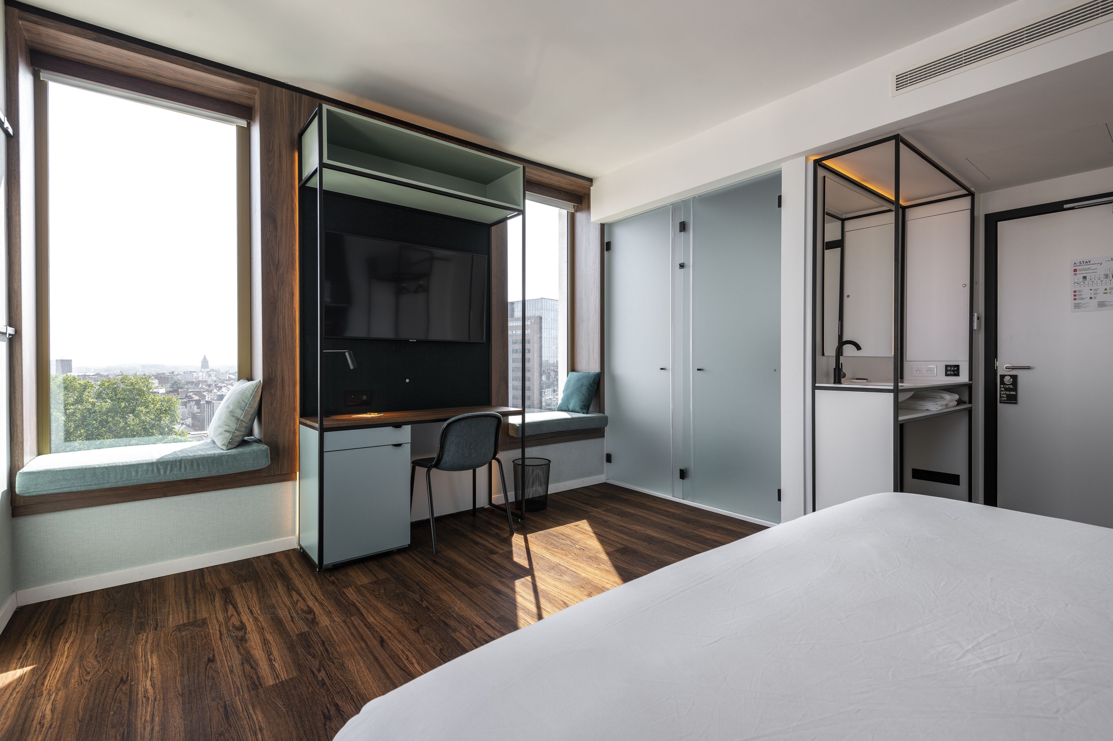 A modern hotel room with a large bed, a desk with a chair, a built-in TV, and a window seat with a view of the city.