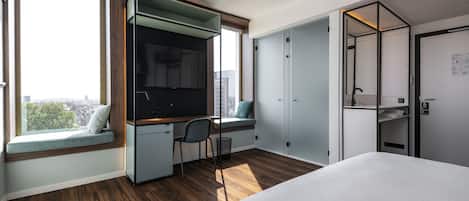Una habitación de hotel moderna con una cama grande, un escritorio con silla, televisión integrada y un asiento junto a la ventana con vistas a la ciudad.
