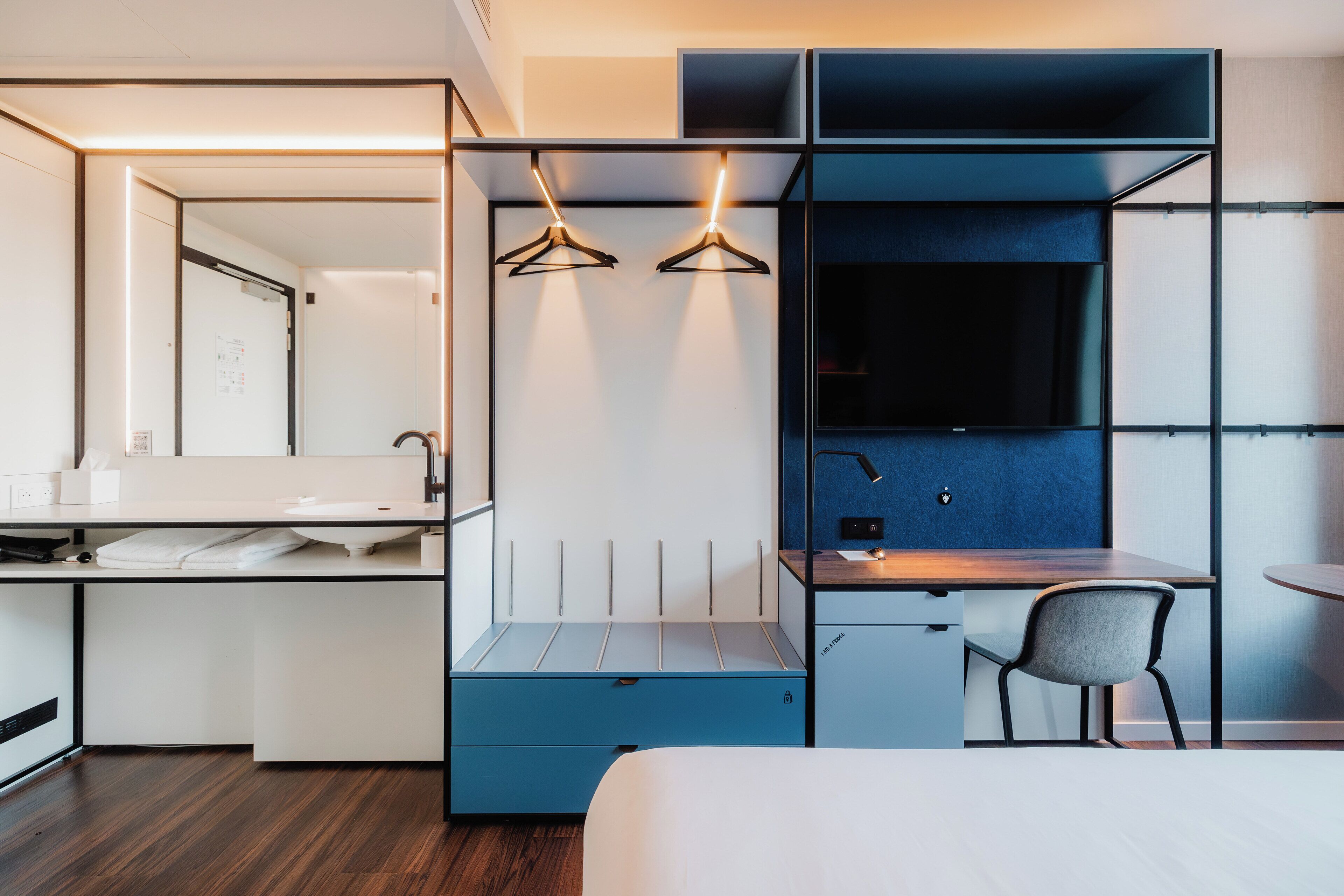 A modern hotel room with a bed, a desk with a chair, a wardrobe, and a mirror.