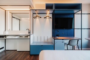 A modern hotel room with a bed, a desk with a chair, a wardrobe, and a mirror.