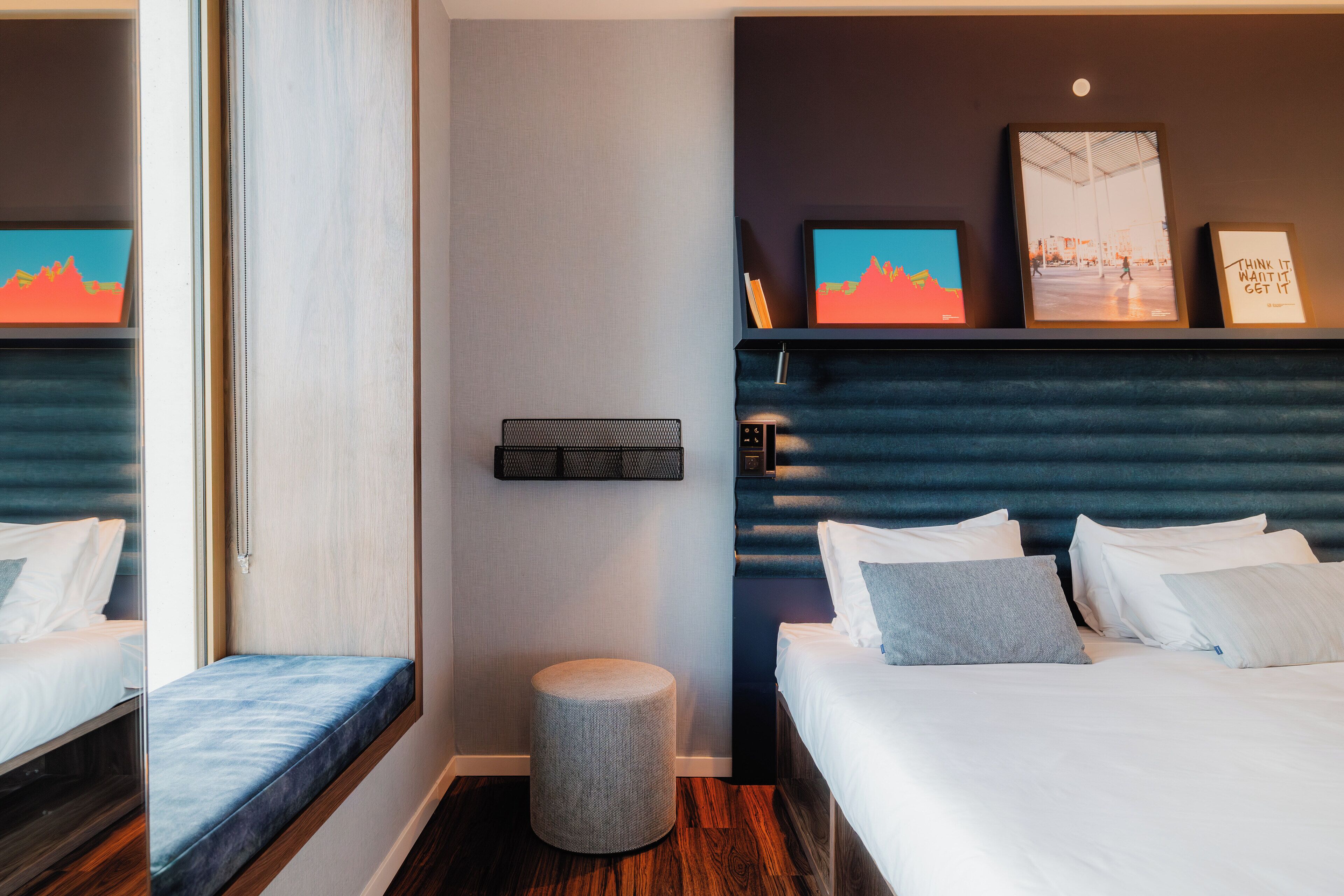 Ein Hotelzimmer mit Bett, Nachttisch und einem kleinen runden Hocker. Darüber befinden sich zwei Bilder an der Wand.