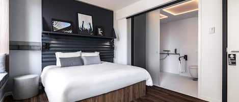 Una habitación de hotel moderna con una cama grande, una banca integrada, un pequeño rincón de descanso, un cabecero oscuro y vistas al baño.