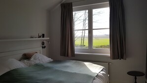 A bedroom with a bed, a window with curtains, a bedside table, and a shelf with items. - De Haas (Oostkapelle)