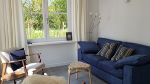 A living room with a blue sofa, a wicker chair, a small table, and a large window with curtains. - De Haas (Oostkapelle)