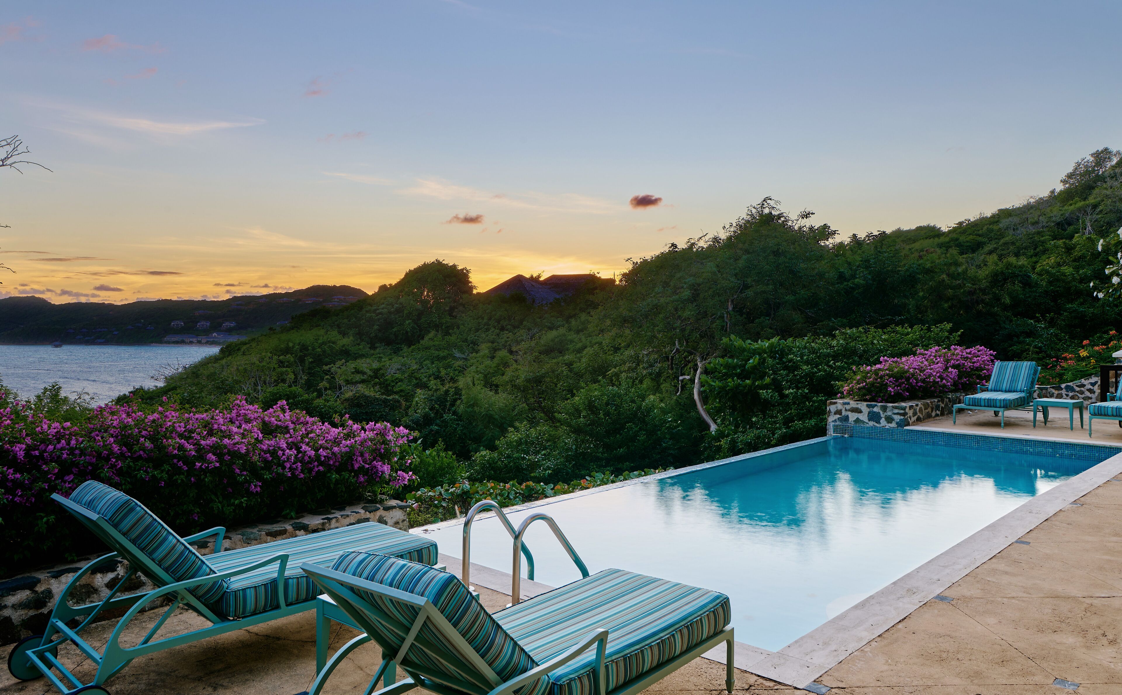 A pool with lounge chairs and a view of the sea at sunset.