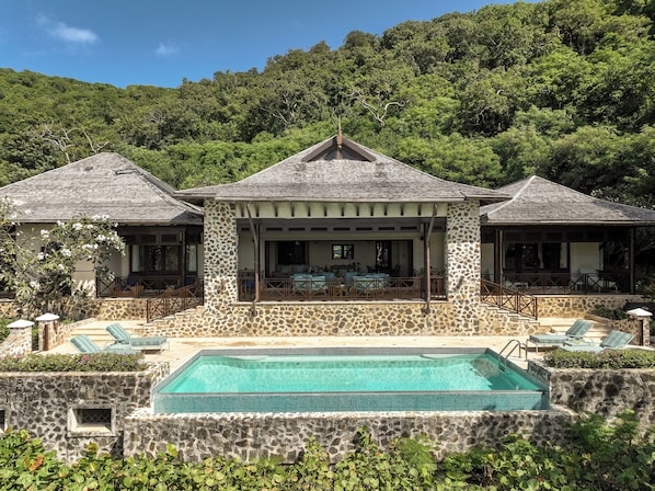 Um edifício de pedra com três andares, piscina, área de estar ao ar livre e vista para uma encosta arborizada.