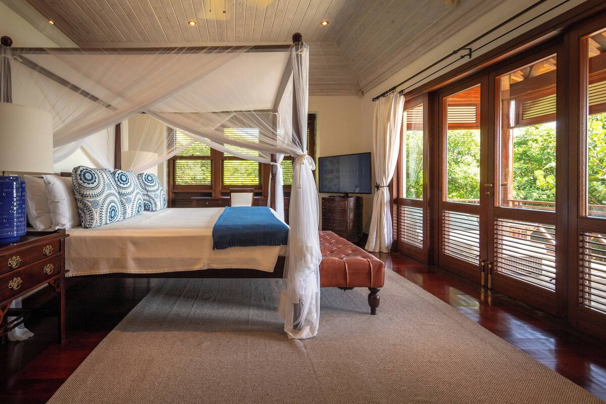 A bedroom with a four-poster bed, a television, and large windows with blinds.