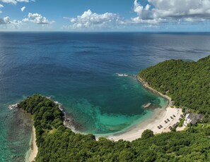 Vista aérea de um resort tropical com águas turquesa cristalinas, colinas verdes exuberantes e uma praia arenosa com guarda-sóis e um edifício com telhado de palha.