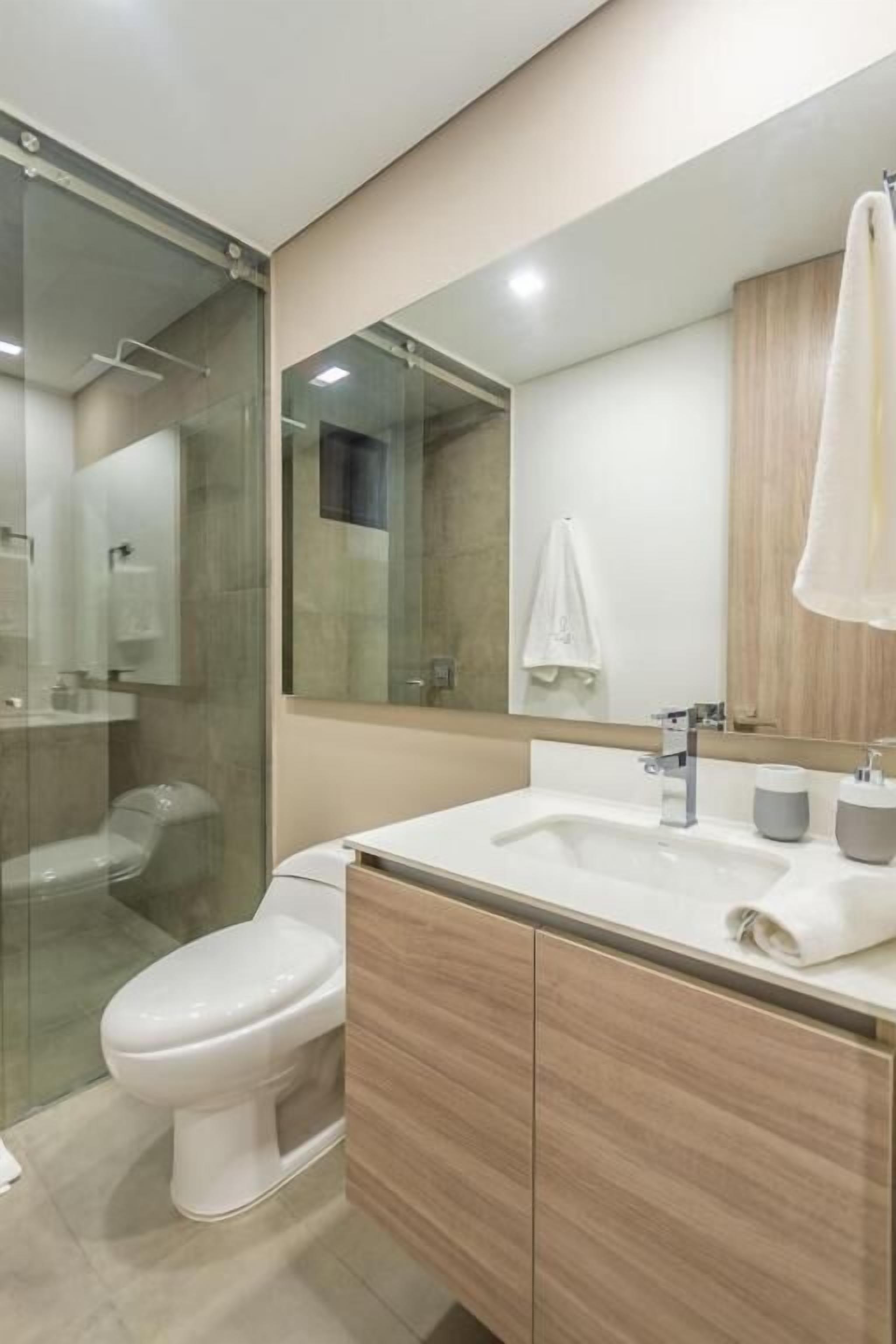 Un baño moderno con cabina de ducha de vidrio, inodoro blanco, tocador color beige con espejo y lavamanos blanco.