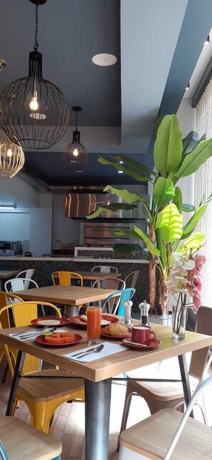 Un comedor moderno con mesas y sillas de madera, una planta grande en maceta y lámparas colgantes.