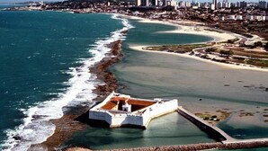 An aerial view of a coastal city with a prominent white fort-like structure near the shore, a wide beach, and a dense urban area with high-rise buildings in the background. - Apartamento Mobiliado Mozart, 105 (Natal)