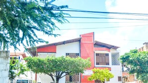 A two-story building with a red and white facade, a tiled roof, and a fenced front yard with trees. - Apartamento Mobiliado Mozart, 105 (Natal)