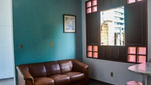 A room with a brown leather sofa, a small round table, and a window with decorative shutters. - Apartamento Mobiliado Mozart, 105 (Natal)