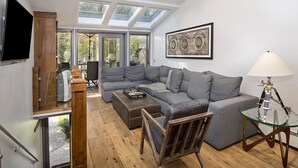 Una sala de estar moderna con un sofá seccional gris, una mesa de centro de vidrio y una silla de madera. Hay un televisor montado en la pared y una lámpara sobre una mesa de vidrio.