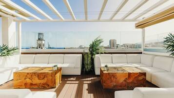 Un área de estar exterior moderna con sofás blancos y mesas de centro de madera, con vistas a un paisaje urbano.