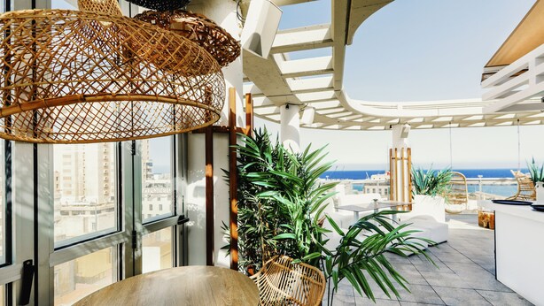 Una terraza moderna al aire libre con vistas a la ciudad y al mar, con mobiliario de mimbre, plantas en macetas y una estructura de pérgola blanca.