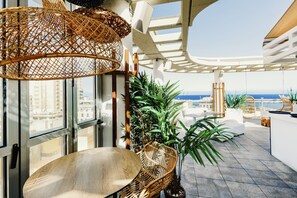 Une terrasse extérieure moderne offrant une vue sur la ville et la mer, aménagée avec des meubles en osier, des plantes en pot et une pergola blanche.