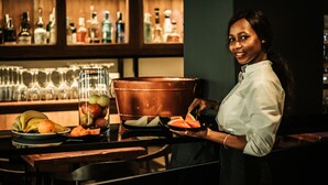 Zona de bar con un bartender preparando una bebida, una olla de cobre y una presentación de frutas.