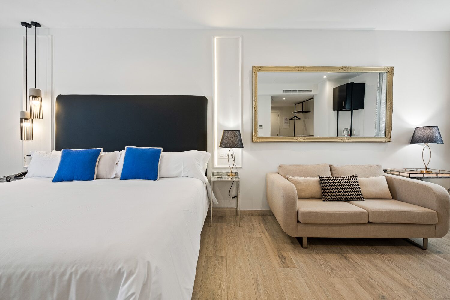 A modern hotel room with a large bed, a sofa, a mirror, and a nightstand with lamps.