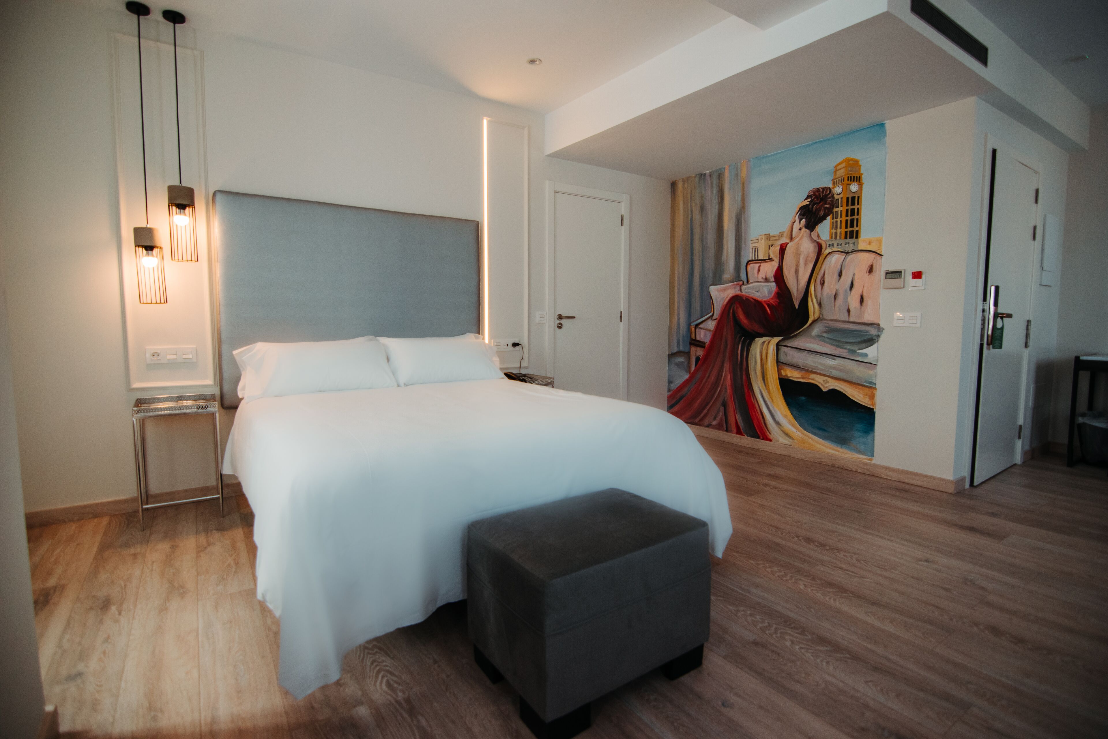 Une chambre d’hôtel moderne avec un grand lit, une table de chevet, un petit pouf et un tableau au mur.