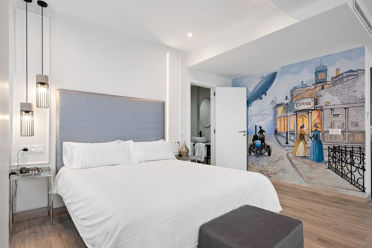 Une chambre d’hôtel moderne avec un grand lit, une table de chevet avec une lampe, une petite table avec un ordinateur portable, et une fresque représentant une scène de rue sur le mur.