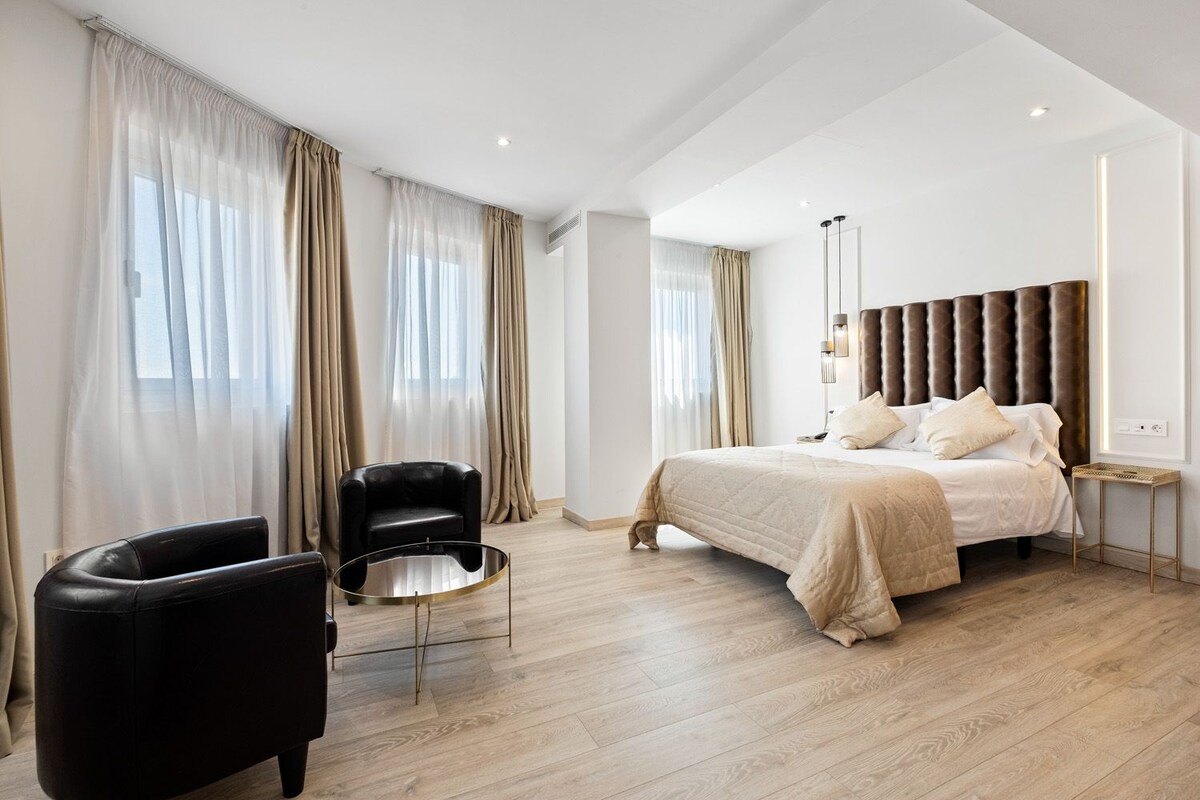 A modern hotel room with a large bed, a black leather chair, a small round table, and sheer curtains.