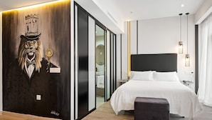 Una habitación de hotel moderna con una cama grande, un pequeño rincón de descanso, una pared con un cuadro de león y una puerta corredera que da a un baño.