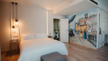 Habitación de hotel con una cama grande, una mesita de noche, un mural en la pared y un baño visible a través de una puerta abierta.